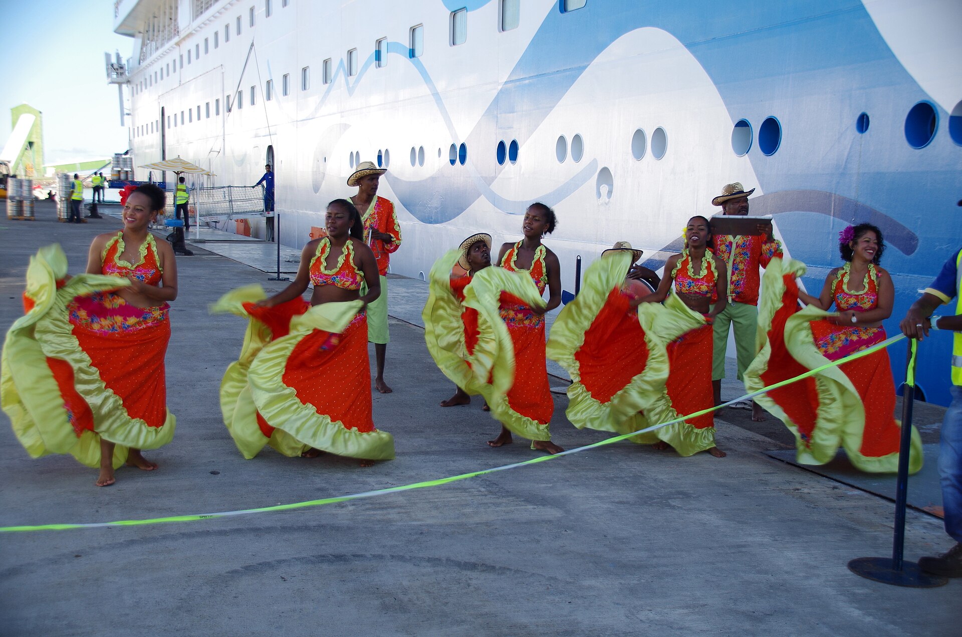 Sega dancers in orange and yellow dresses with musicians beside a cruise ship in Mauritius
